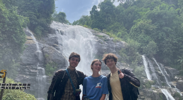 three people in their 20s, one female standing between two males, with a waterfall and green trees behind them
